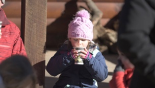 Child in a pink hat and blue coat sits outdoors, drinking hot cocoa from a cup.