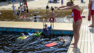 A swim instructor on a dock demonstrates to a group of children in the water at a beach. Other people are swimming and enjoying the background.