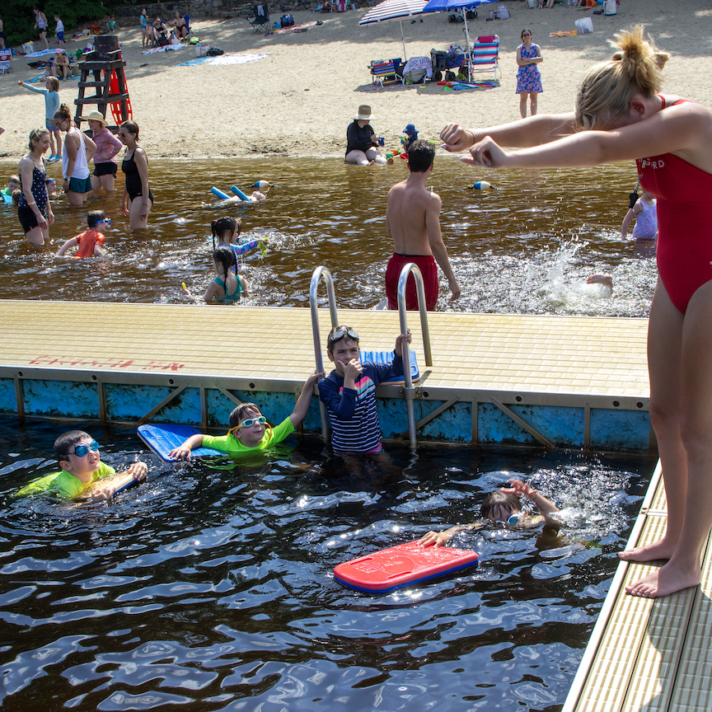 A swim instructor on a dock demonstrates to a group of children in the water at a beach. Other people are swimming and enjoying the background.