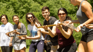 A group of people outdoors participate in a team-building activity, balancing a marble along wooden tracks.