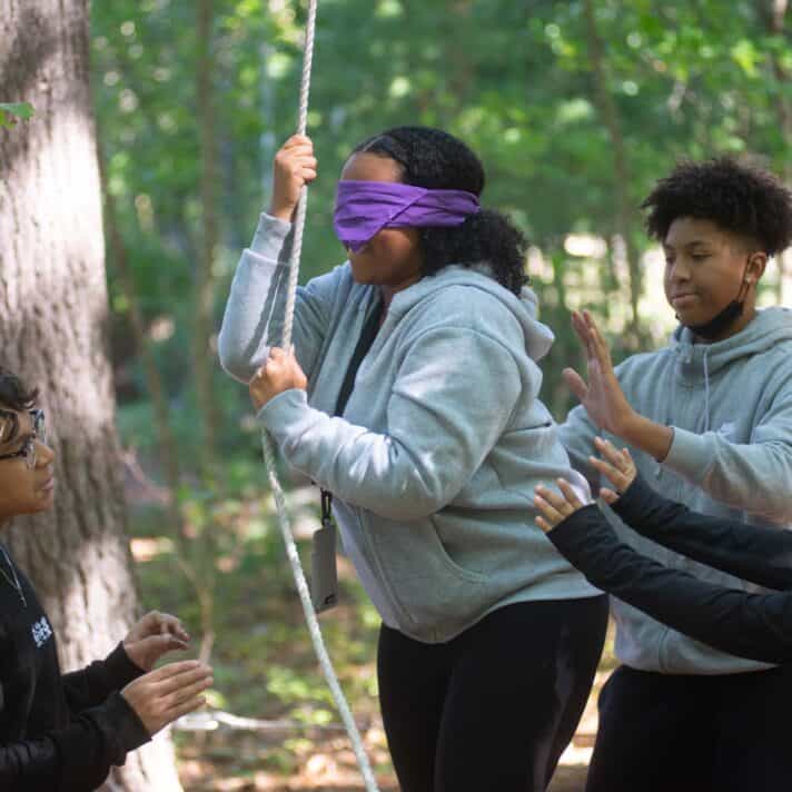 Blindfolded teenager holding rope helped by other campers.