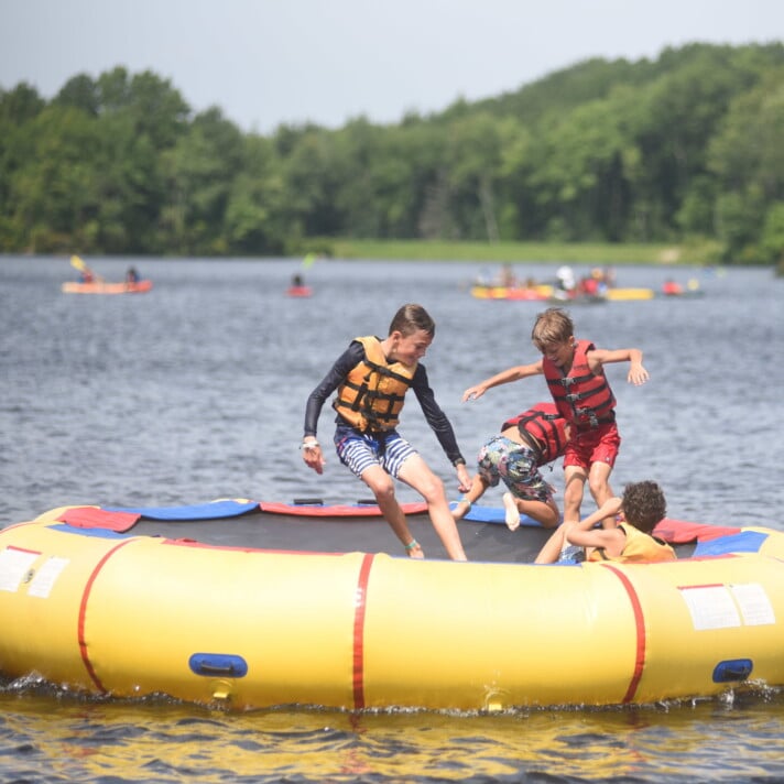 A group of kids playing on an inflatable raft in a pond.