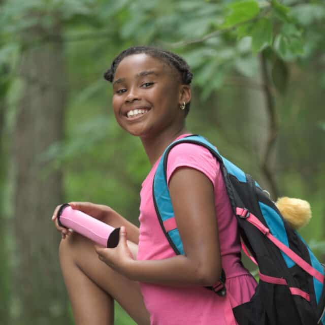 A young girl in a pink backpack is sitting on a rock in the woods.