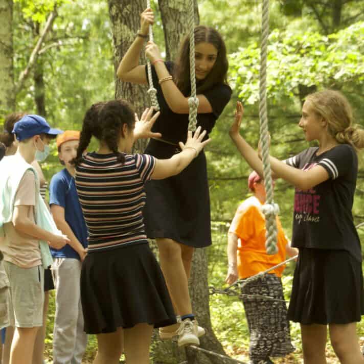 Students help each other balance on a suspended tightrope on a ropes course.