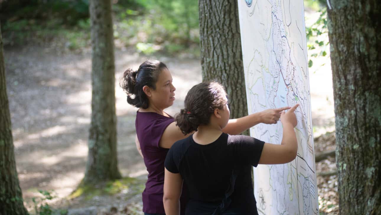Two women standing next to a map in the woods.