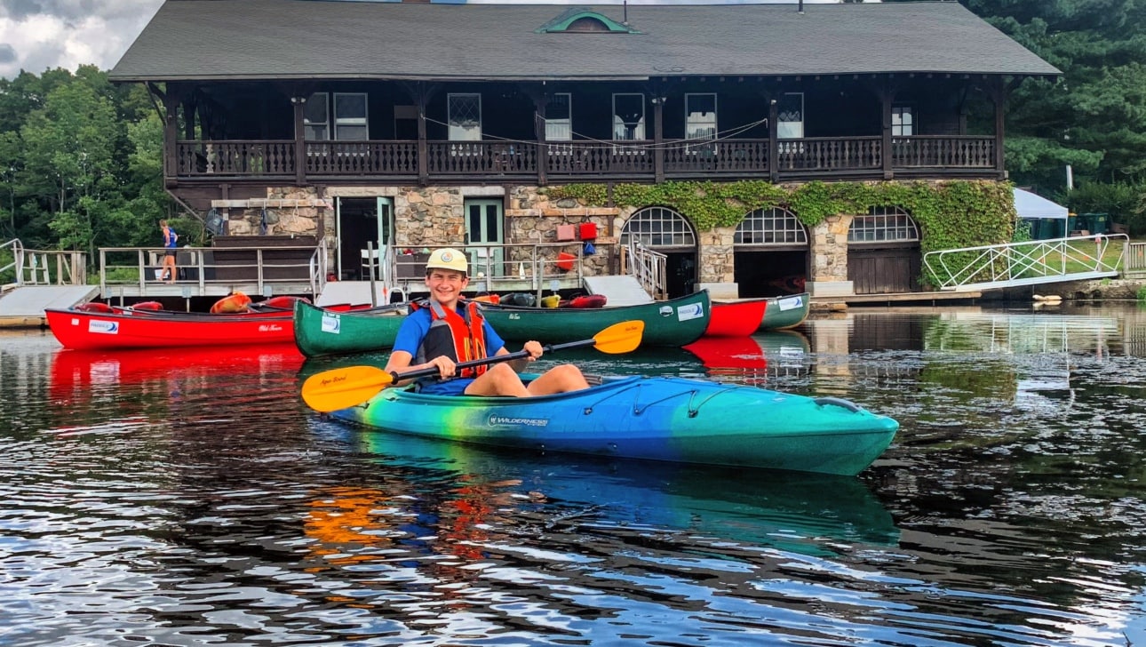 A teenager paddling a kayak in front of a boathouse.