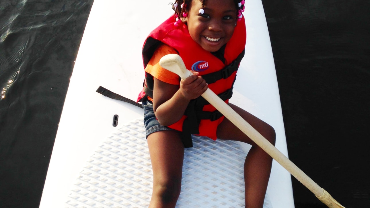A young girl sits on a stand-up paddbleboard (SUP) in a river.