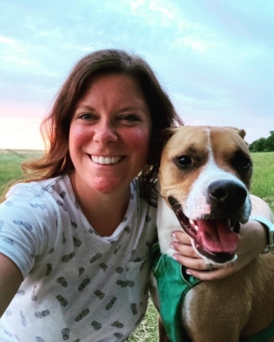 A woman smiling and a dog panting in a field at sunset