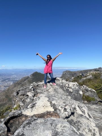 A woman standing on the top of a mountain smiling at the camera and raising her hands in victory