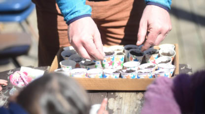 Counselor puts small paper cups with dirt and seeds onto tray, while children watch during outdoor Earth Day Activity at spring camp week.