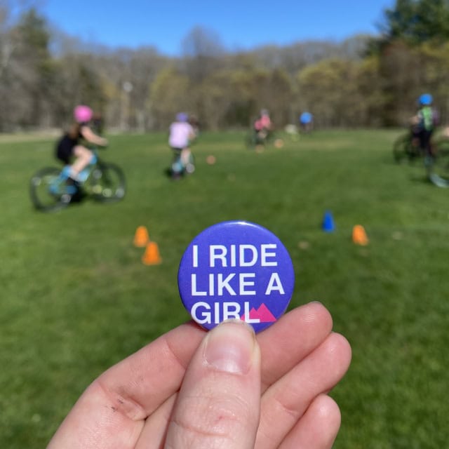 Close-up of a hand holding a button that says "I RIDE LIKE A GIRL." In the background, people are cycling on a grassy field with scattered cones.