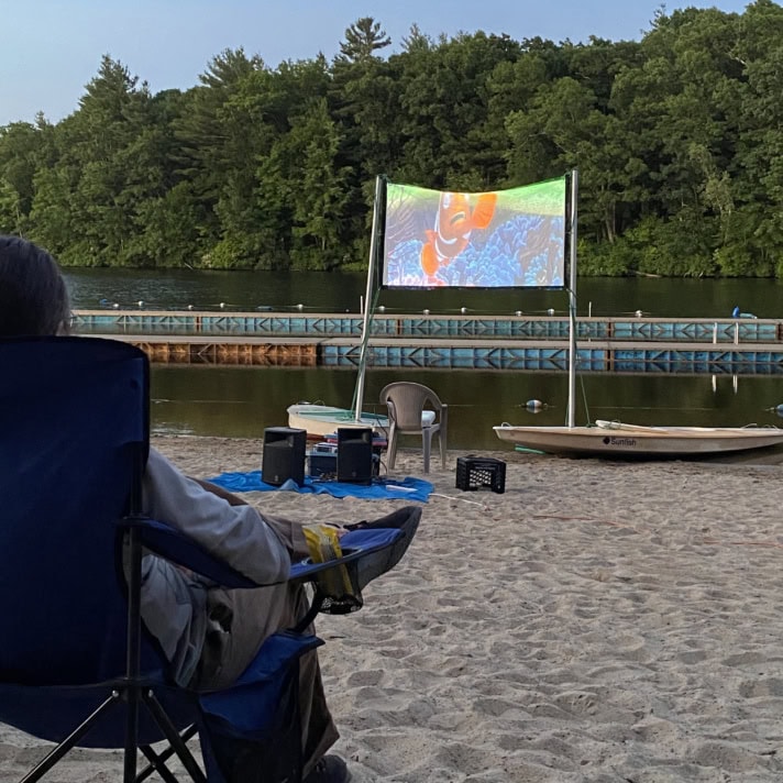 A person sits in a camp chair on a sandy beach watching an outdoor movie projected on a screen by a lake, with trees and kayaks in the background.
