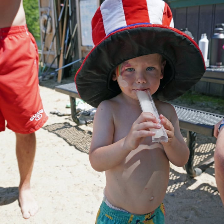 A young child wearing a large patriotic hat eats an ice pop while standing shirtless outdoors near a bench on a sunny day.