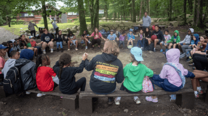 A group of campers and counselors join hands in a circle in the woods.