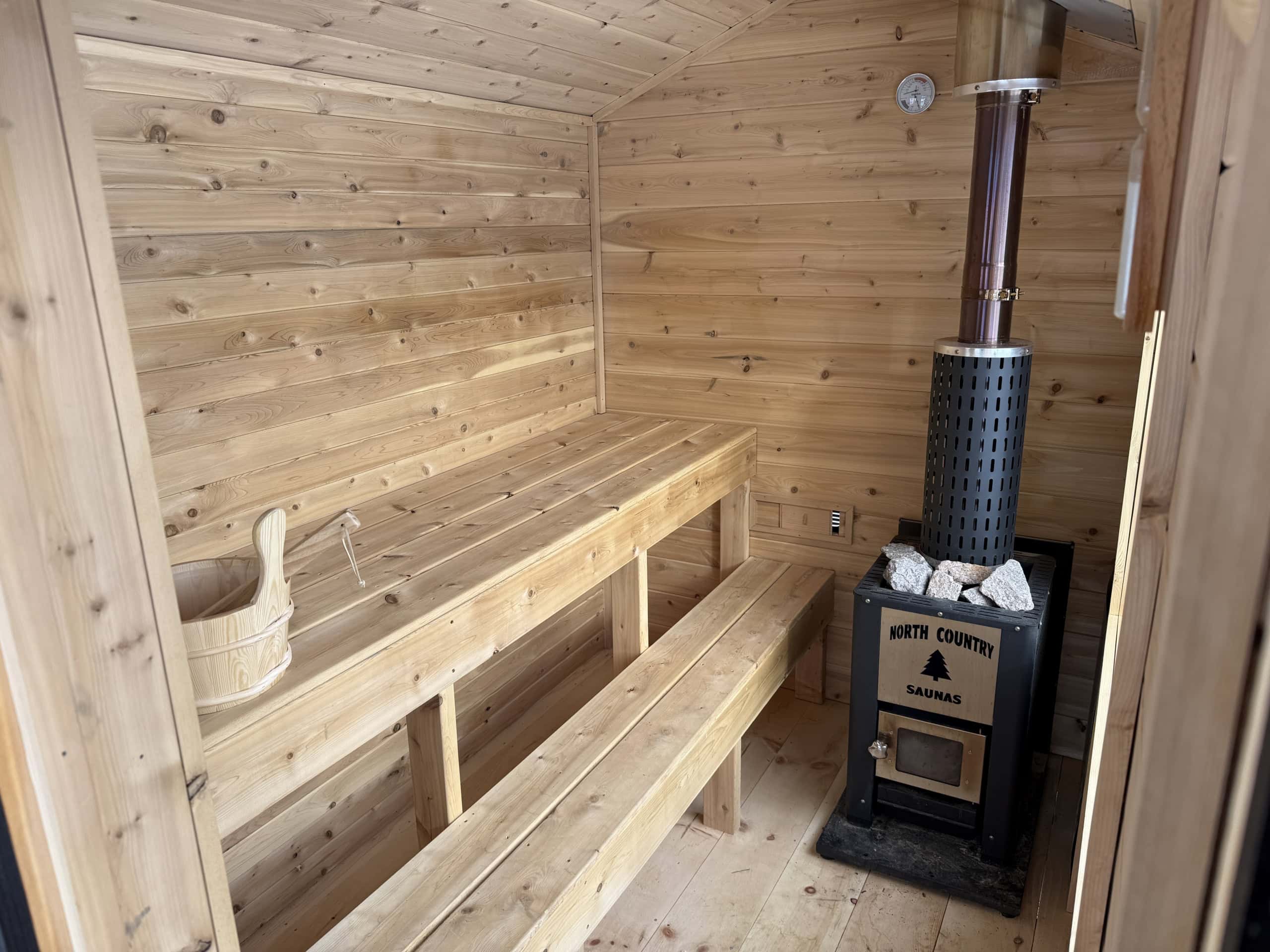 A wooden sauna interior with two benches, a bucket and ladle on the left, and a wood-burning stove on the right.