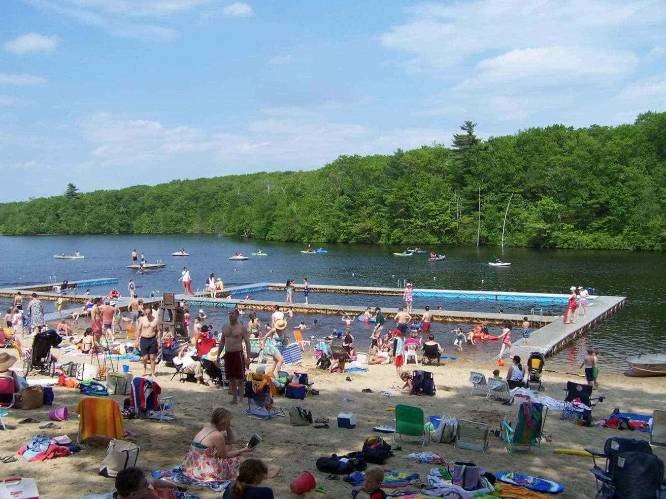 A crowded lakeside beach with people swimming, sunbathing, and kayaking. A wooden dock extends into the water, surrounded by a forested shoreline under a blue sky with clouds.
