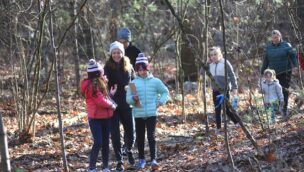 A group of people, including children and adults, walk through a forest in winter clothing. The ground is covered with dry leaves, and bare trees surround them.