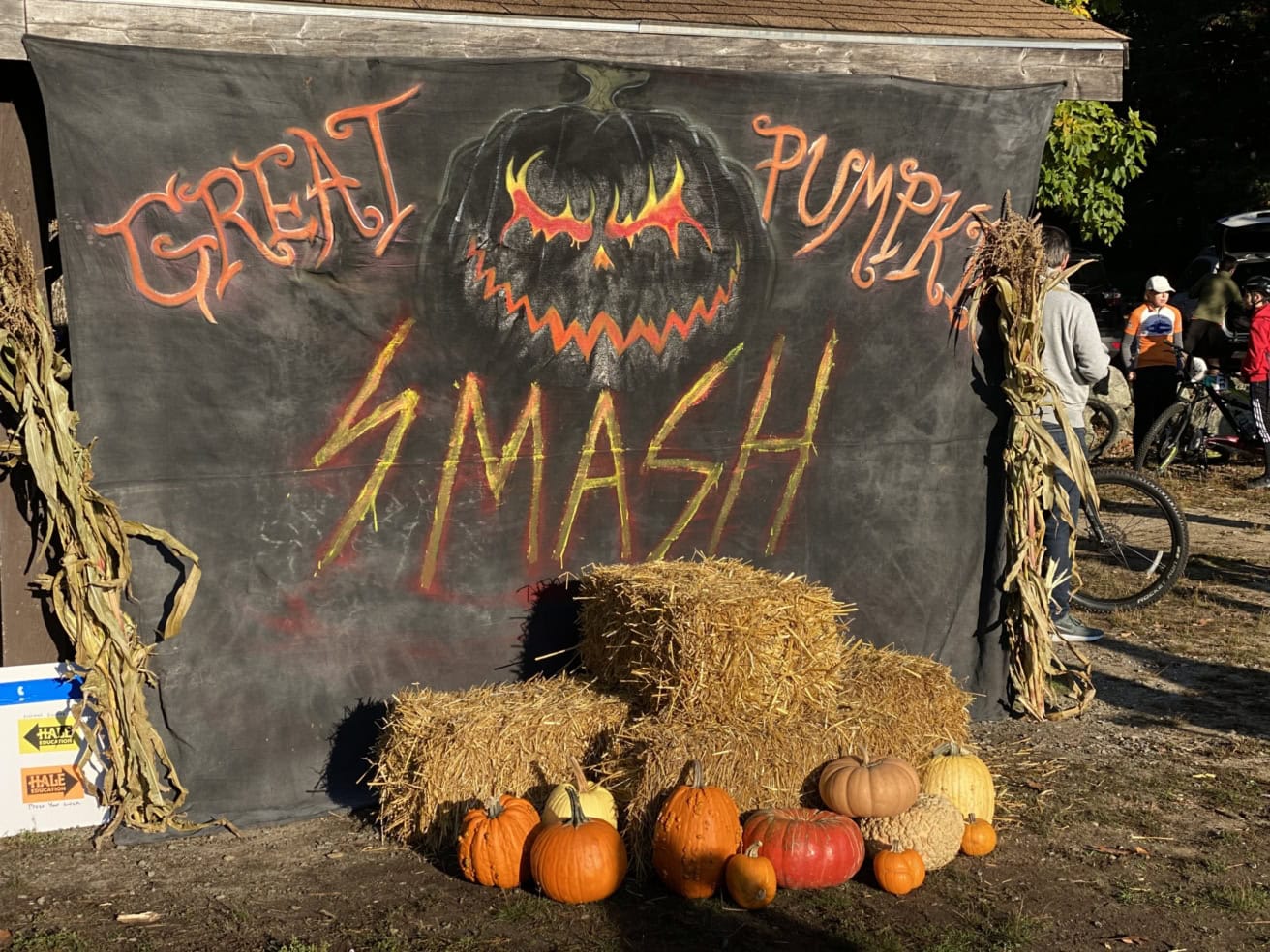 A sign with a menacing pumpkin face reads "Great Pumpkin Smash." Around it are hay bales and various pumpkins, with people and bikes in the background.