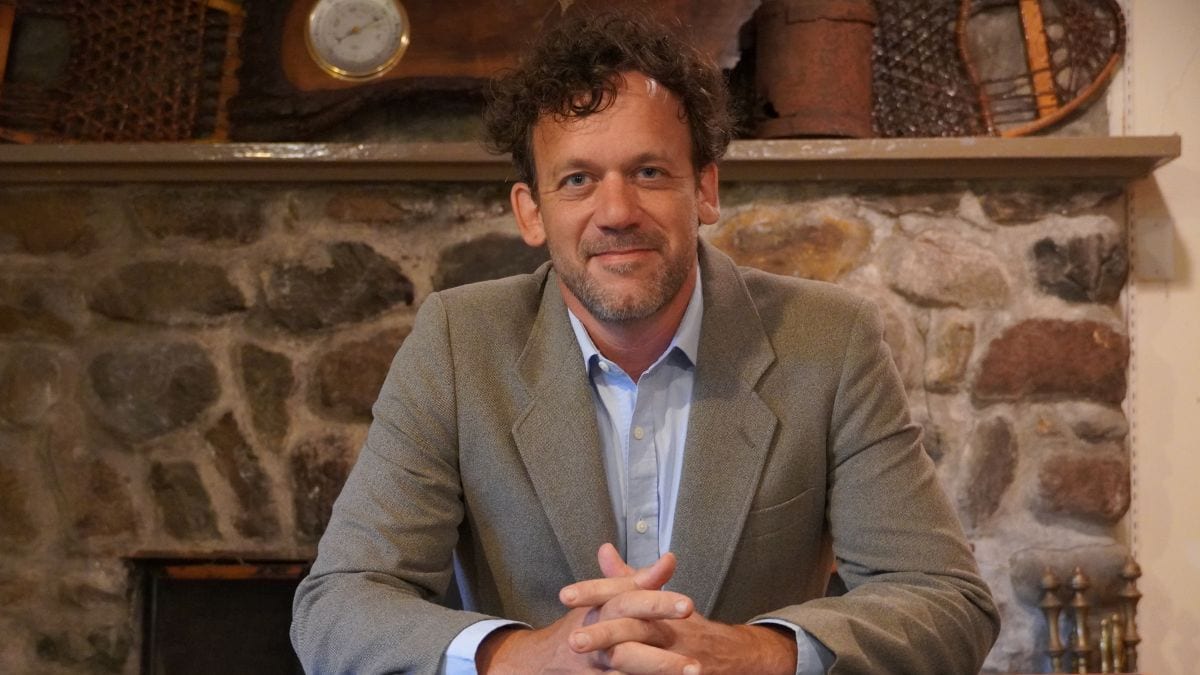 A man with curly hair and a beard, wearing a gray blazer and blue shirt, sits in front of a stone fireplace with rustic decor.