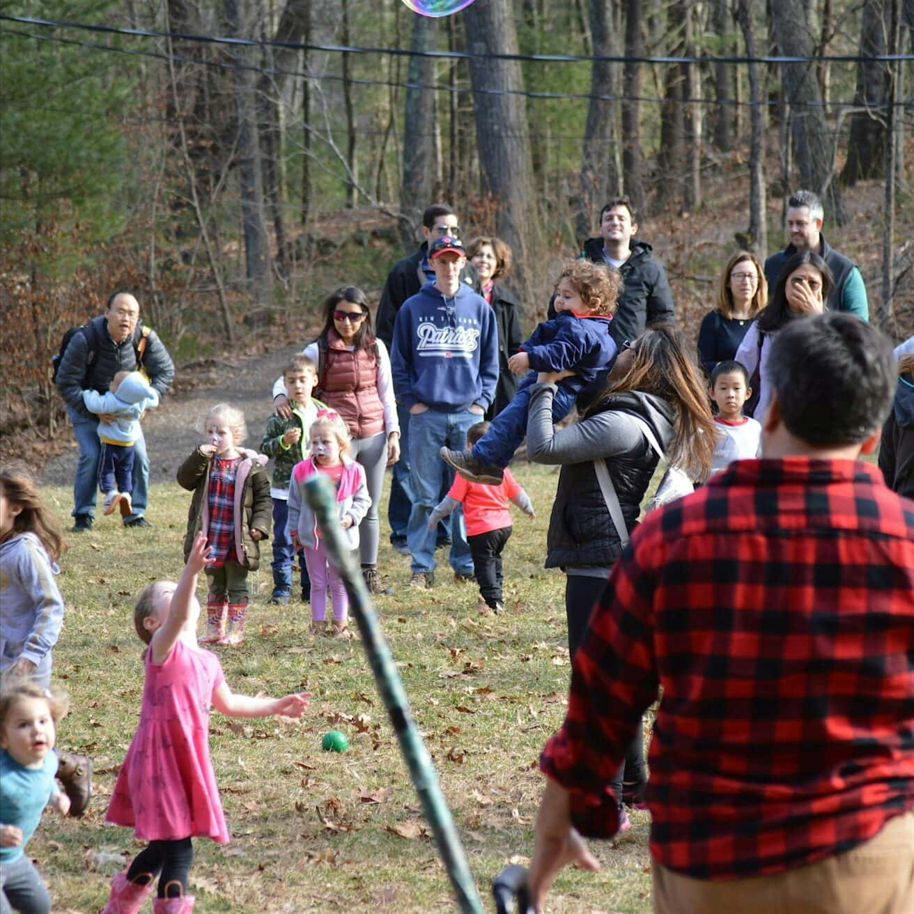 A group of people, including adults and children, gather outdoors on grass with trees in the background. Some children play with a bubble.