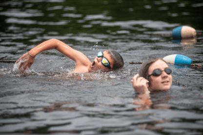 Two children wearing goggles swim in an outdoor body of water, with one doing a freestyle stroke and the other partially submerged near floating lane markers.