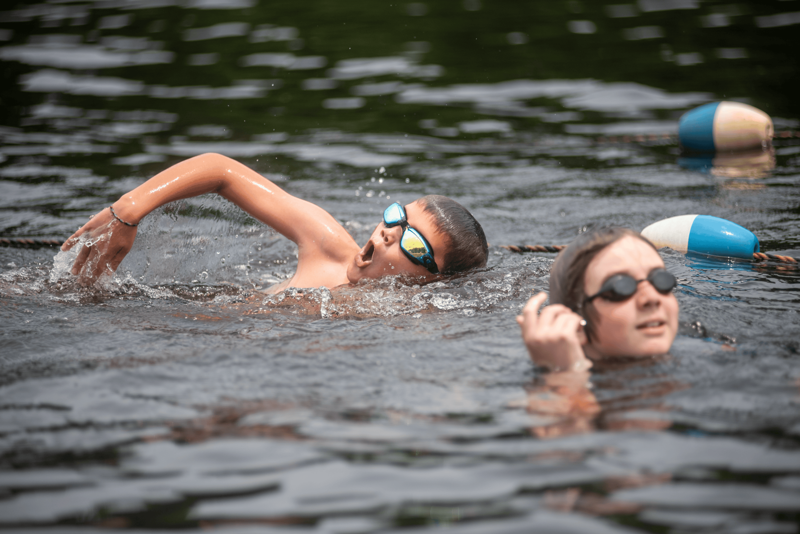 Two children wearing goggles swim in an outdoor body of water, with one doing a freestyle stroke and the other partially submerged near floating lane markers.