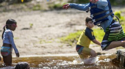 Child in a blue life vest jumps into a lake, while other kids play in the water. Colorful triangular flags are strung in the background.