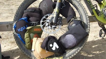 Bicycle wheel leaned against a fence, displaying a variety of knee and elbow pads arranged around the tire.