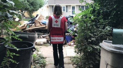 A person wearing a Red Cross disaster relief vest walks toward a pile of debris and trash outside a house.