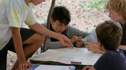 Four boys sit at a wooden table outdoors studying a map. One boy points at the map while others listen. A clipboard and compass are on the table. Trees and leaves are in the background.