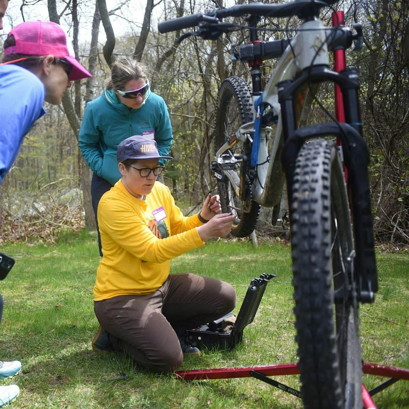Three people gather around a bicycle on a stand. One person kneels, adjusting the bike, while the others watch. They are outdoors on grass with trees in the background.