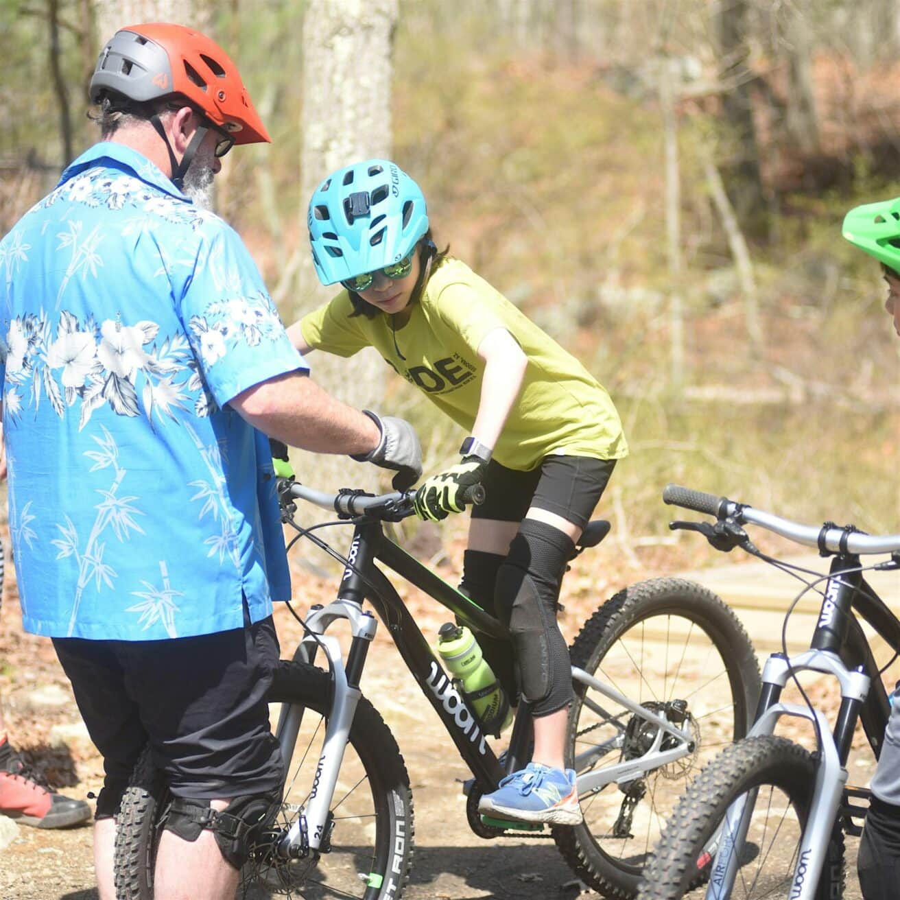 A person wearing a helmet receives instructions while sitting on a bike, guided by another person also wearing a helmet. Another bike is nearby.