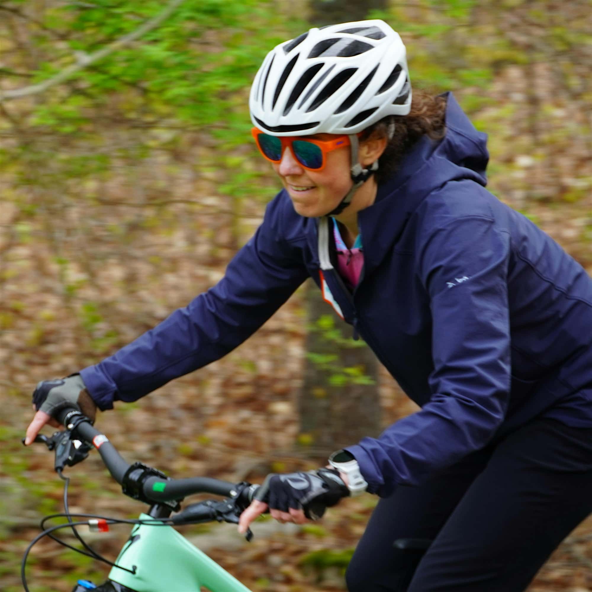 Person cycling outdoors wearing a helmet, sunglasses, and gloves, against a blurred background of greenery and brown leaves.