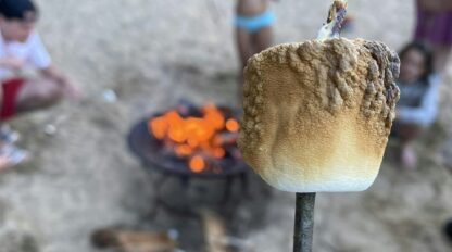 Close-up of a toasted marshmallow on a stick in front of a campfire, with children playing in the background at a sandy beach.