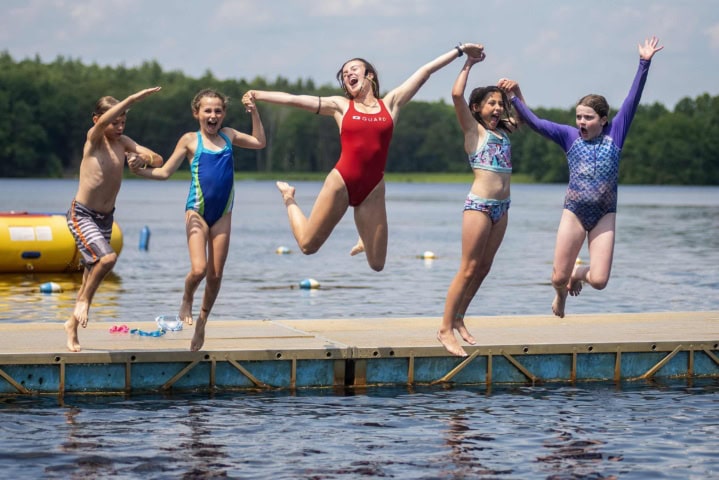 Five children and an adult in swimsuits joyfully jump off a dock into a lake on a sunny day.