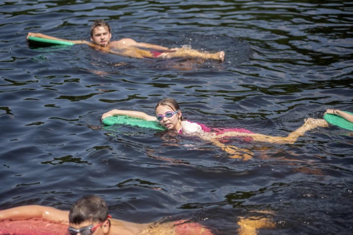 Three people float in a lake, each on a board. The central person wears goggles and a swimsuit, looking at the camera. The other two focus on swimming.