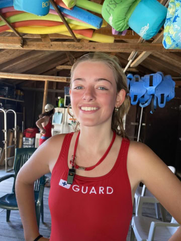 Person wearing a red lifeguard swimsuit stands in a wooden structure with colorful floatation devices in the background.