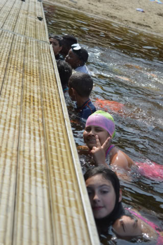 Children swimming near a wooden dock. One child in a pink swim cap is smiling and making a hand gesture.