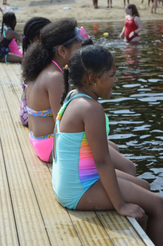 Three children in swimsuits sit on a wooden dock by the water, with people swimming nearby.