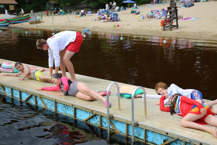 Lifeguard instructs four children in swimsuits on a dock beside a lake, with a sandy beach and people in the background.