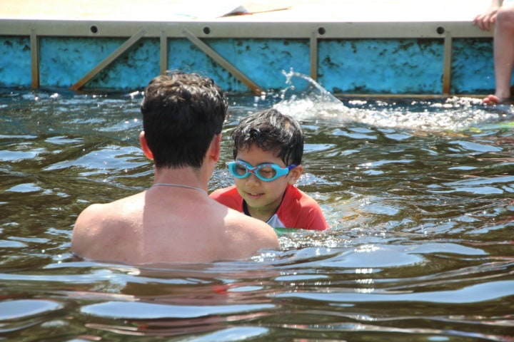 An adult and a child wearing goggles are swimming in a pool. The child is wearing a red top and both are partially submerged in water.