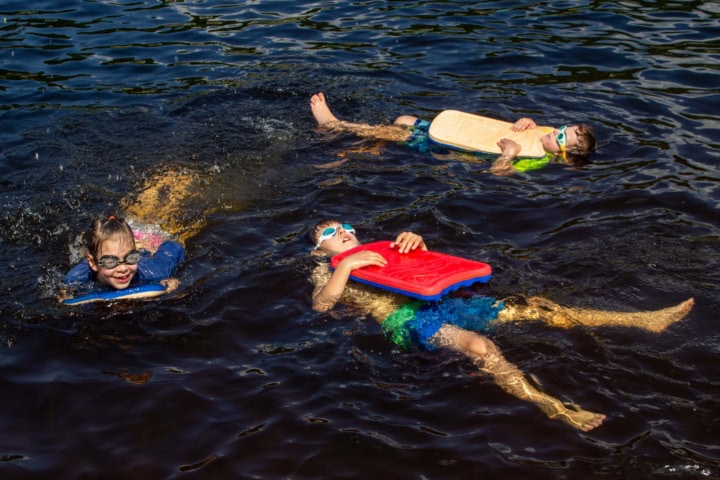 Three children swim in dark water using kickboards and wearing goggles, two float on backs, one swims forward.