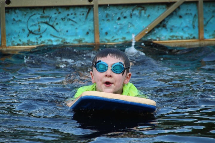 Child wearing goggles swimming with a kickboard in a pool.