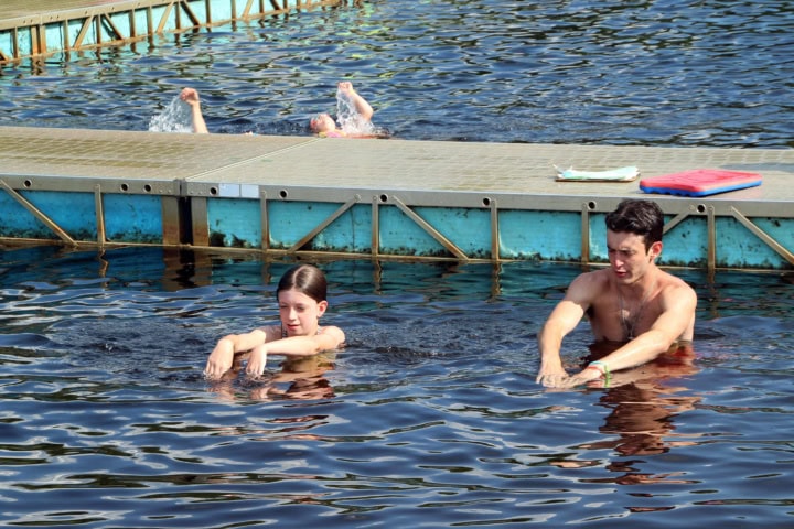 Two people practice swimming strokes in a body of water near a platform, while another swims backstroke in the background.