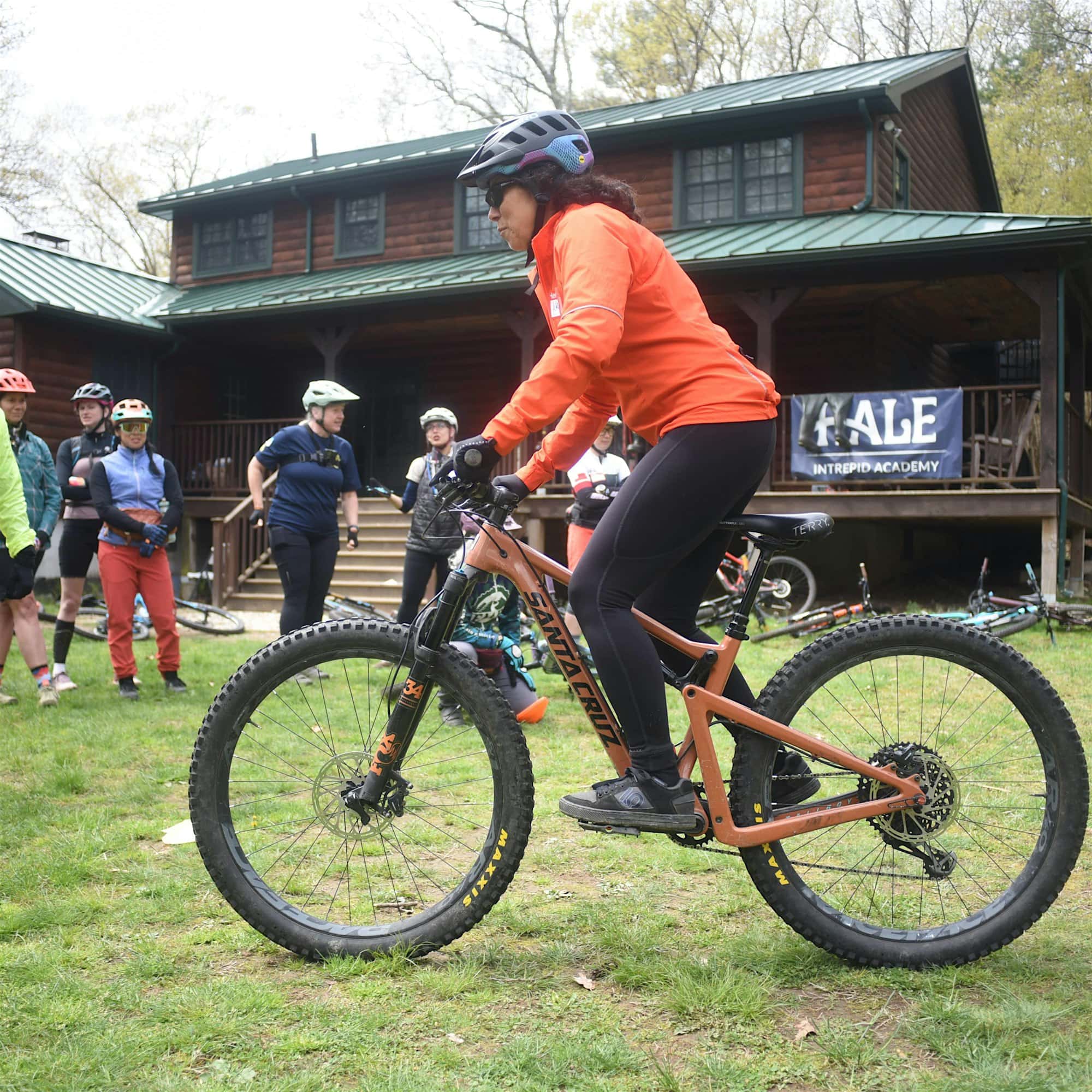 Person in an orange jacket riding a mountain bike on grass, with people watching in front of a wooden building.