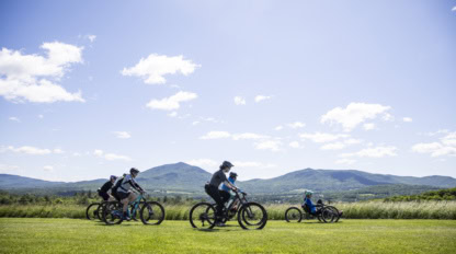 Cyclists ride on a grassy field with mountains and a blue sky in the background.