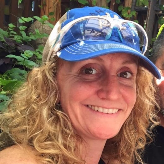 Leslie Riding smiling, wearing a blue cap with sunglasses on top, surrounded by greenery.