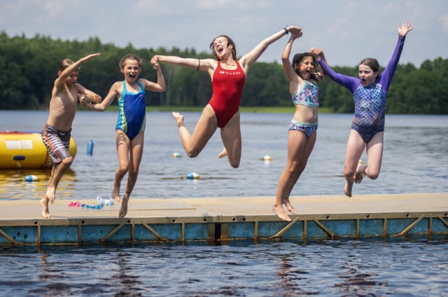 Four kids holding hands with a lifeguard are jumping off a dock. They are smiling and laughing and there are trees in the horizon behind the water's edge.