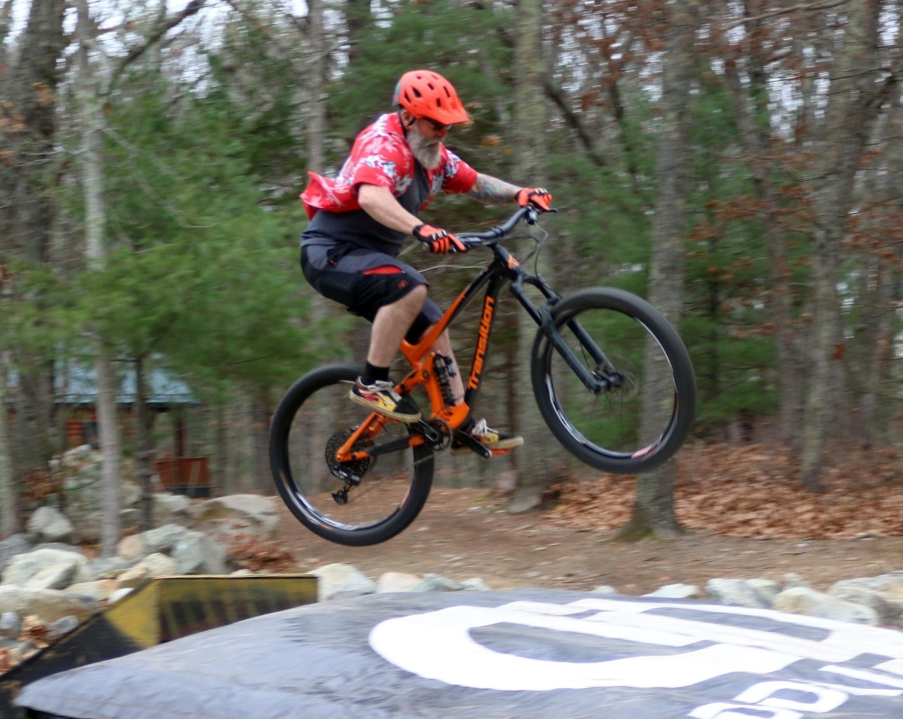 A person wearing a helmet and red shirt is riding a mountain bike in mid-air above an inflatable landing pad in a wooded area.