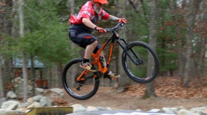 A person wearing a helmet and red shirt is riding a mountain bike in mid-air above an inflatable landing pad in a wooded area.